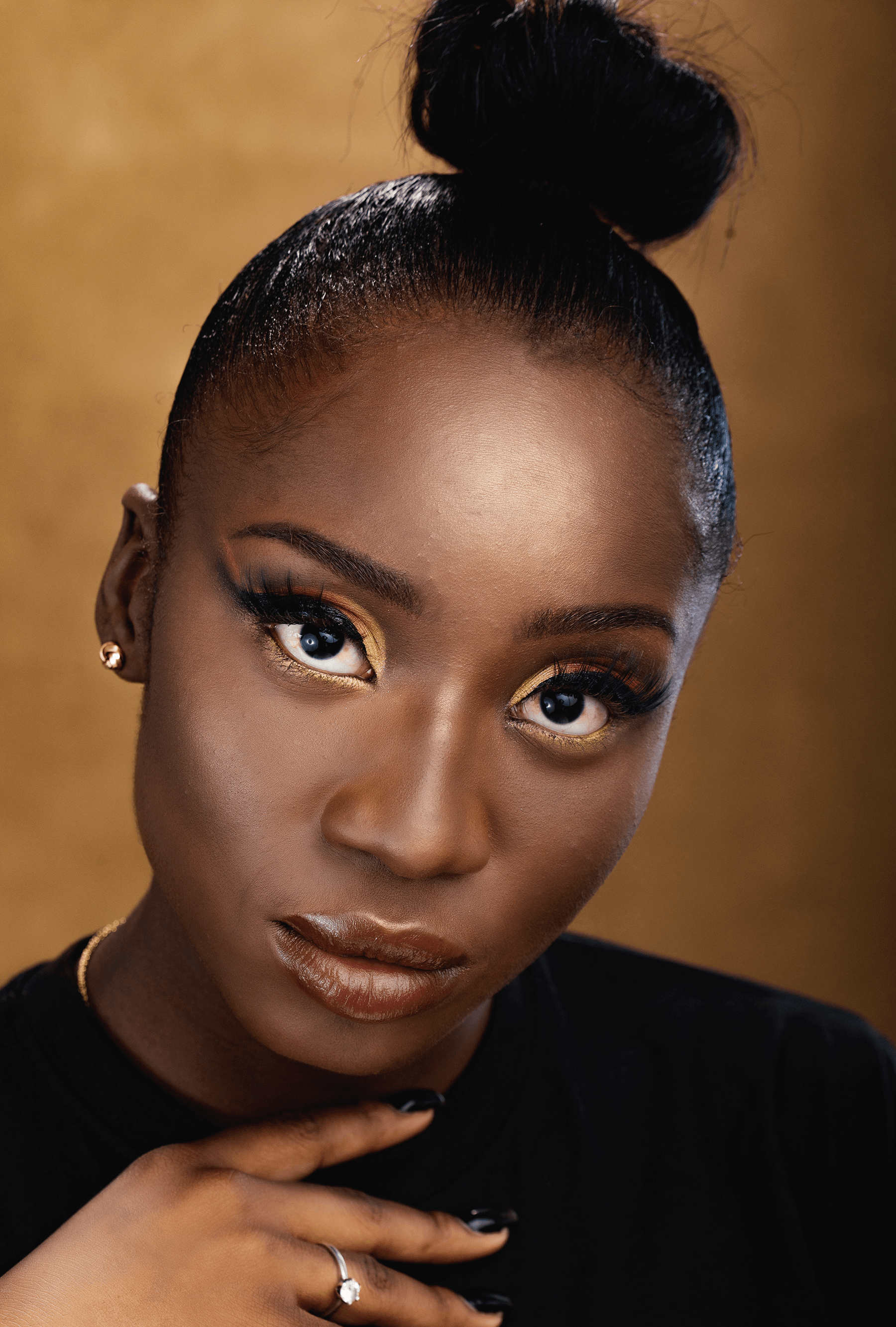 Woman with dark skin, updo, gold eyeshadow, looking at the camera. Black top, ring, gold background.