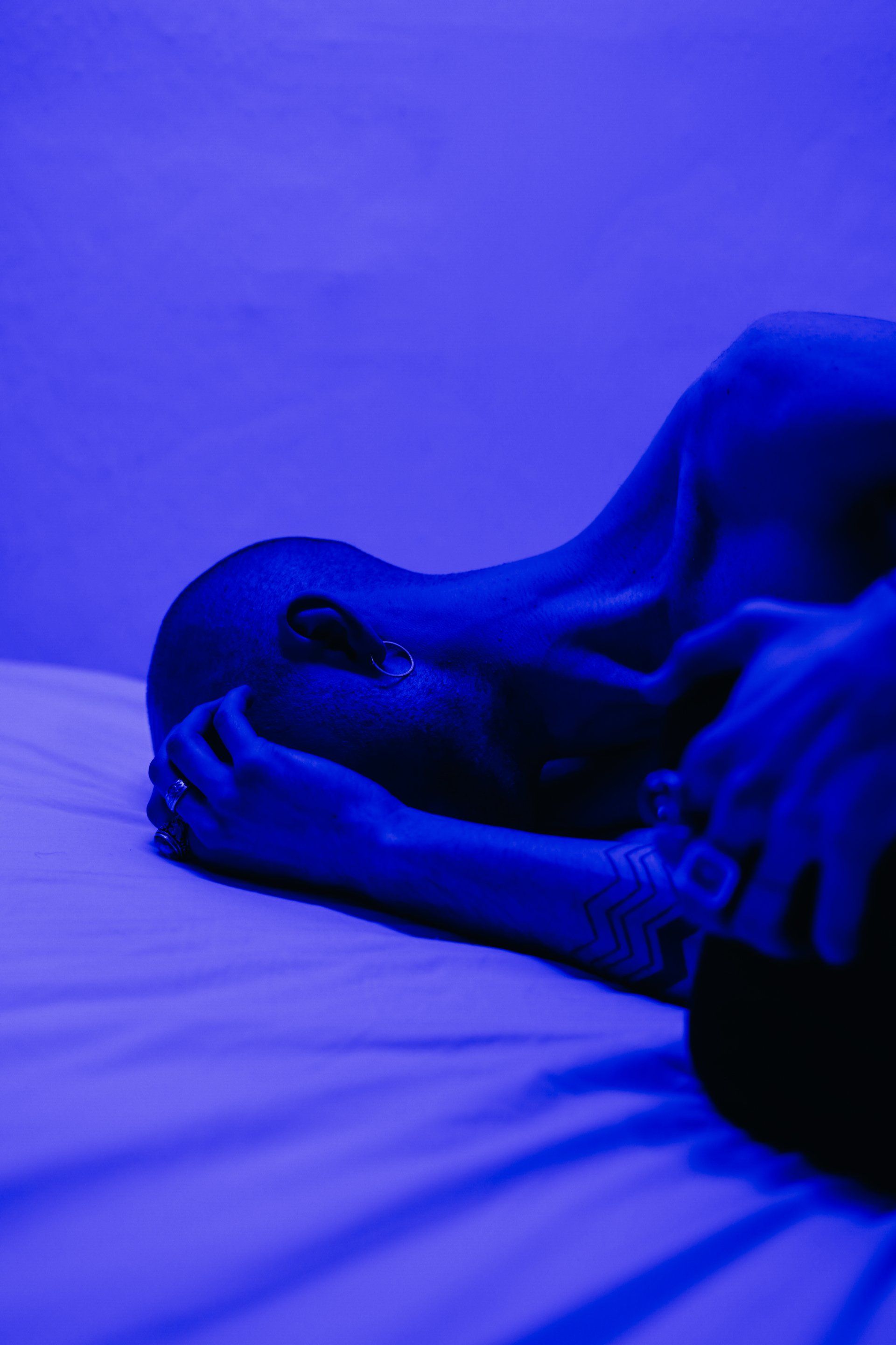 Person lying down, face in hands, illuminated by blue light on a bed.