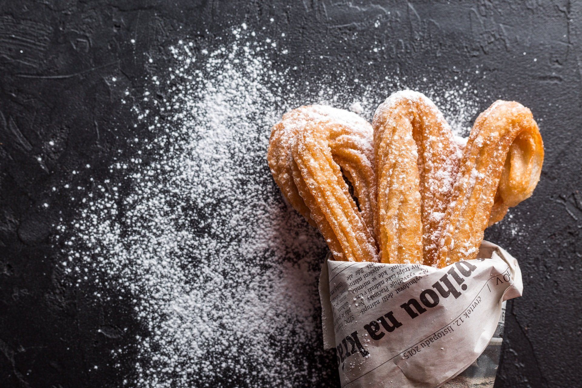 Churros are covered in powdered sugar in a newspaper bag.