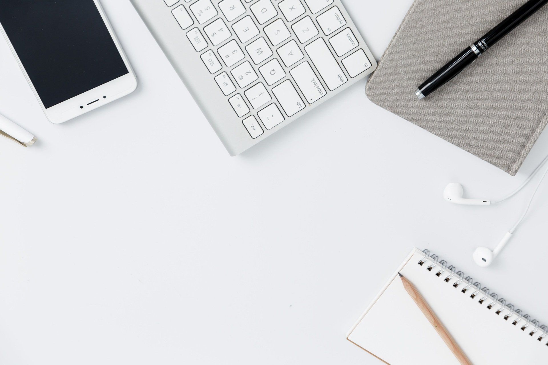 White desk workspace with phone, keyboard, notebook, pen, earbuds, and pencil.