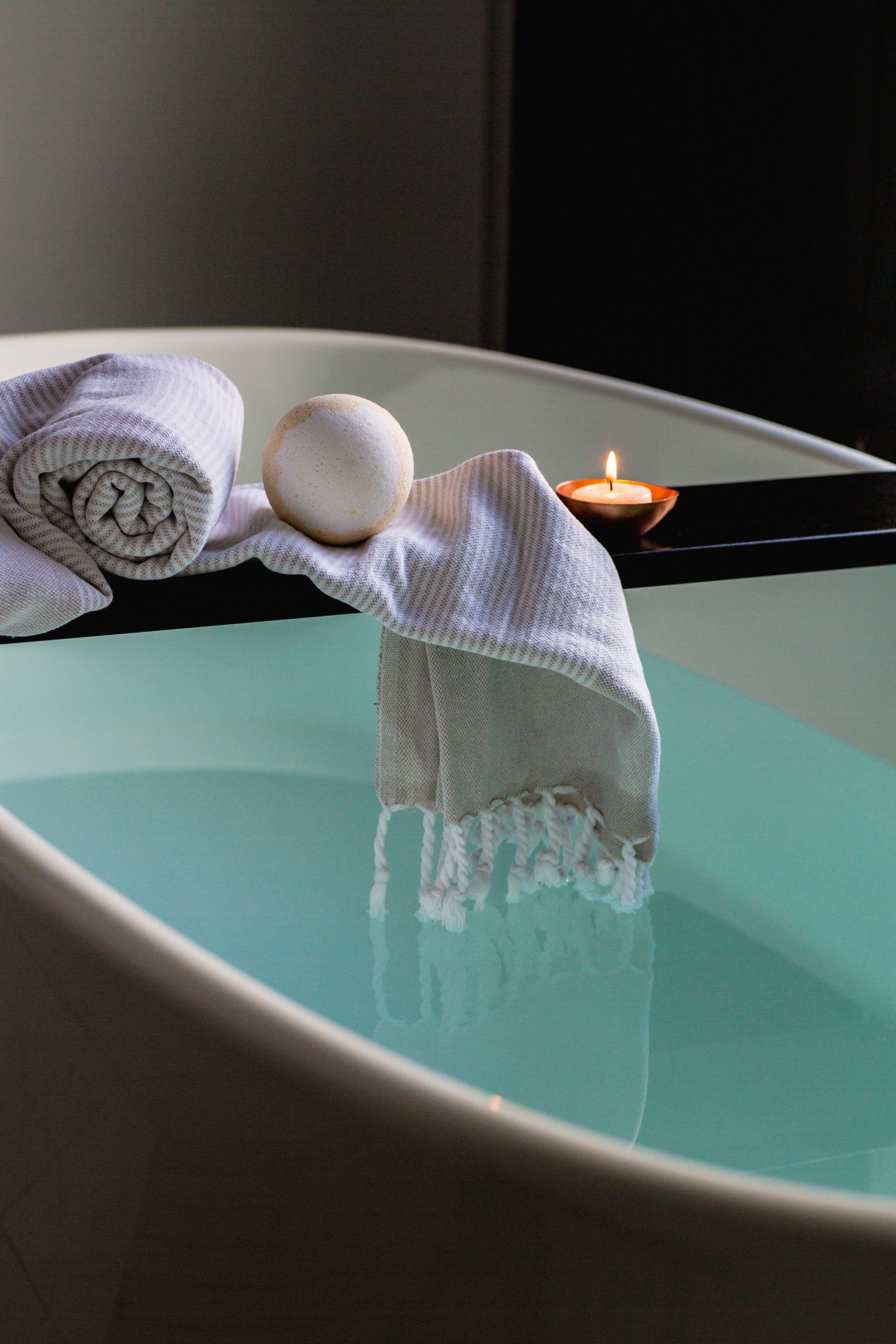 Toalhas, uma bola de spa e uma vela ao lado de uma banheira turquesa.