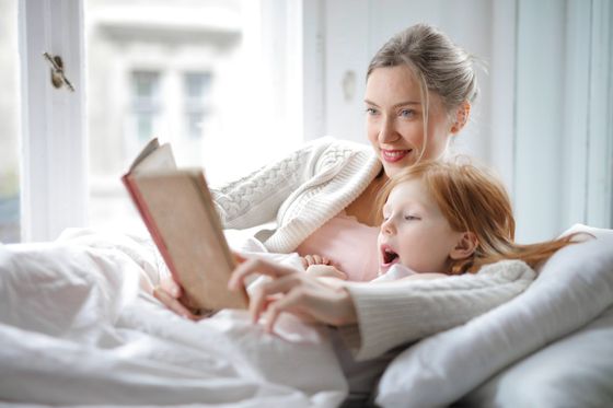Woman reading a book with a young child in bed by a bright window