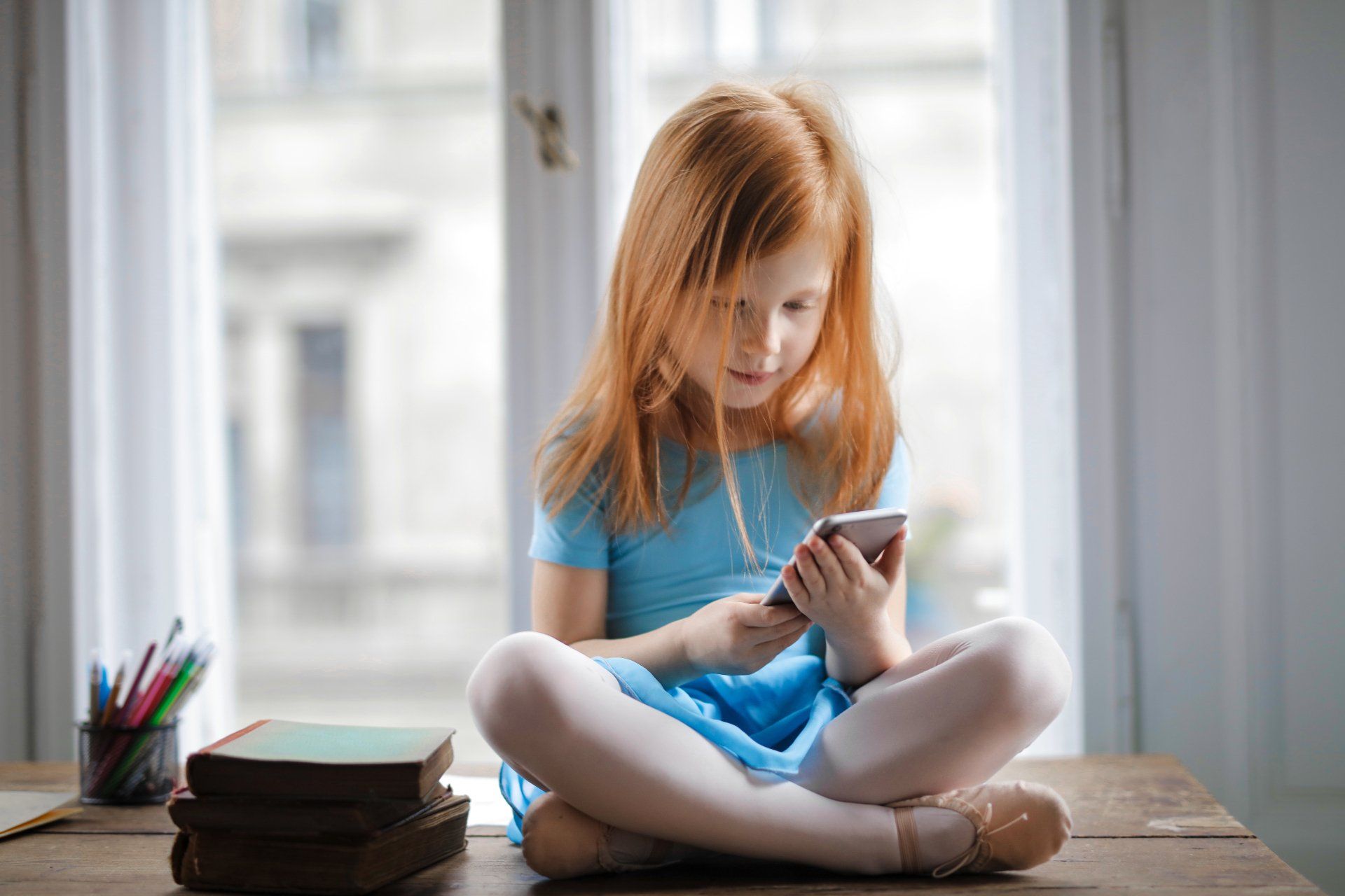 Girl with red hair in a blue dress sits cross-legged, using a smartphone on a table by a window.