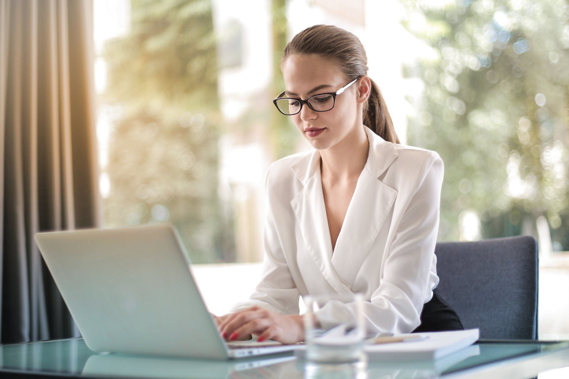 Woman in glasses and white blazer typing on a laptop at a desk.