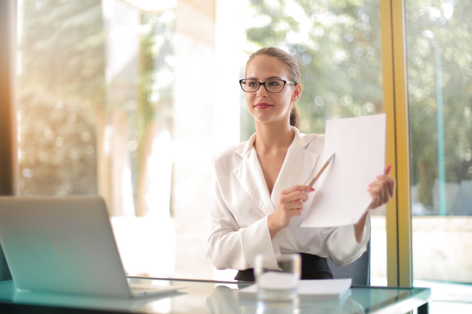 A woman is sitting at a desk with a laptop and holding a piece of paper.