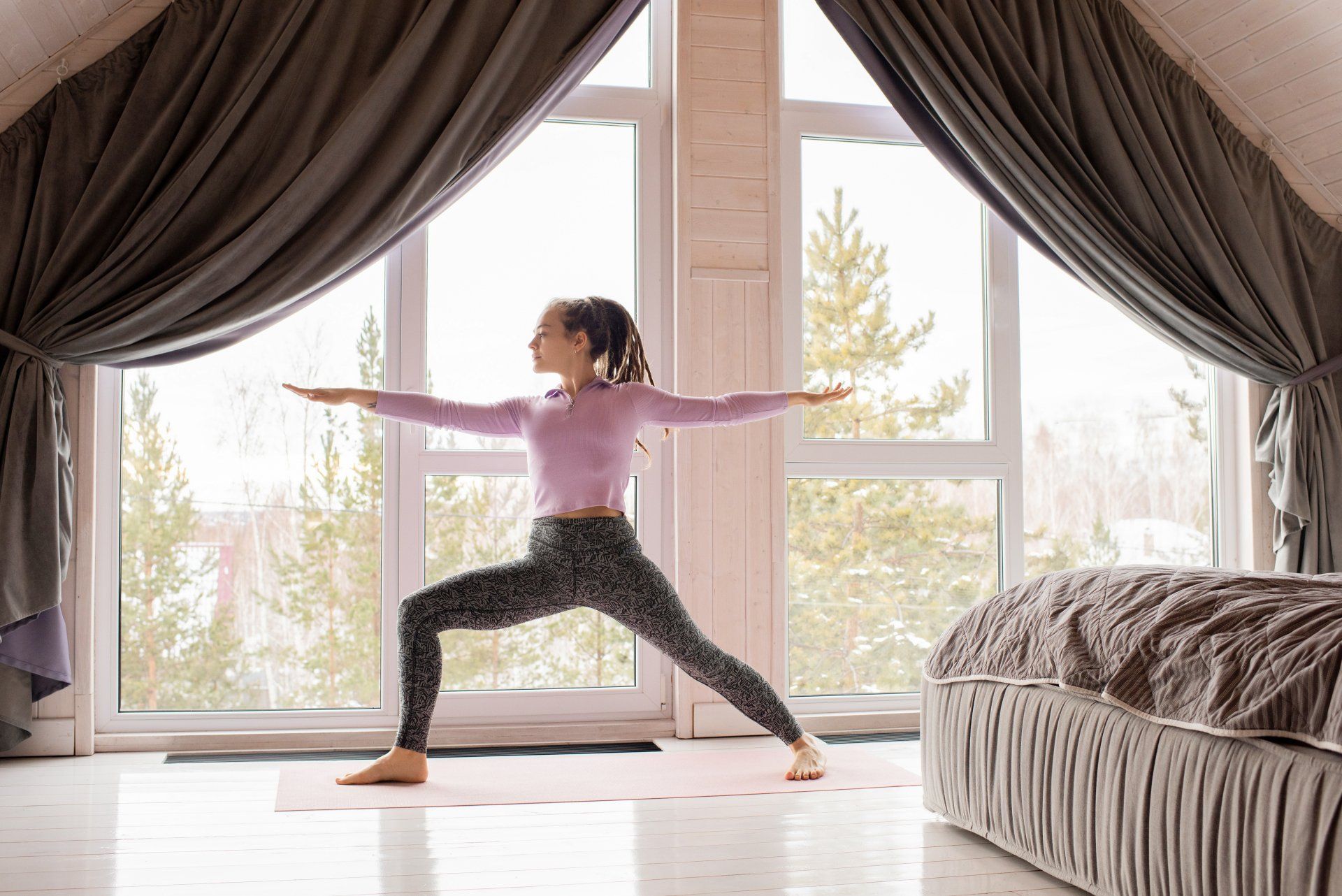 A person in workout clothes performs a yoga warrior pose on a mat in front of large windows with forest views.