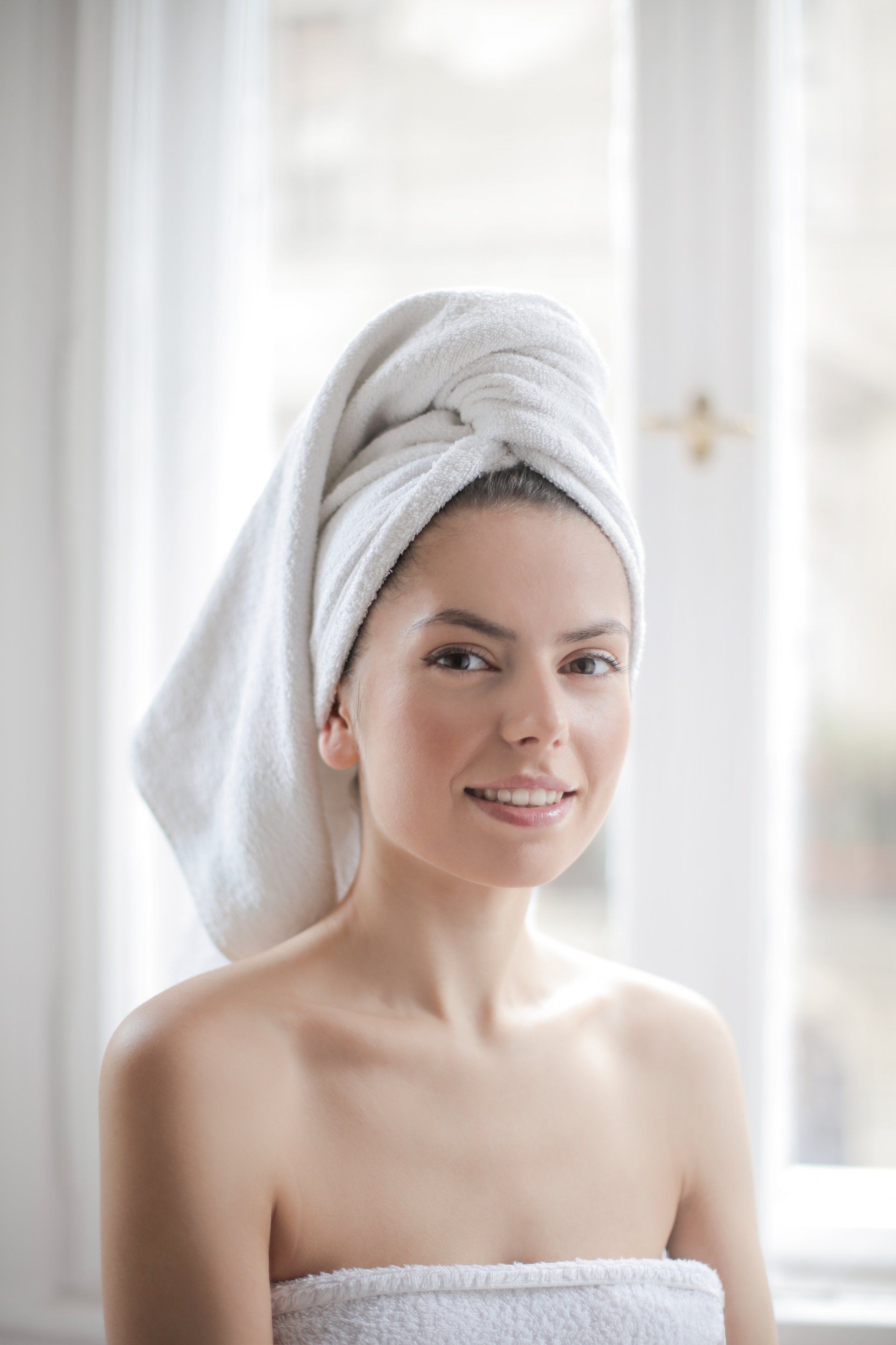 Woman with a white towel wrapped on her head and around her body, smiling. Standing near a window.