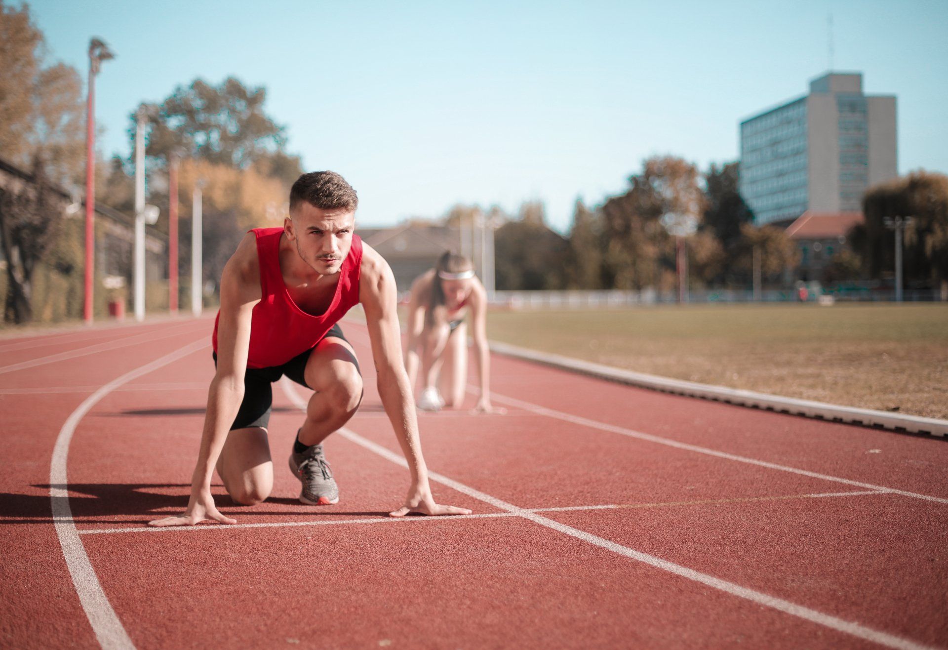 Male runner in red prepares to start, on track with another runner, under clear sky.