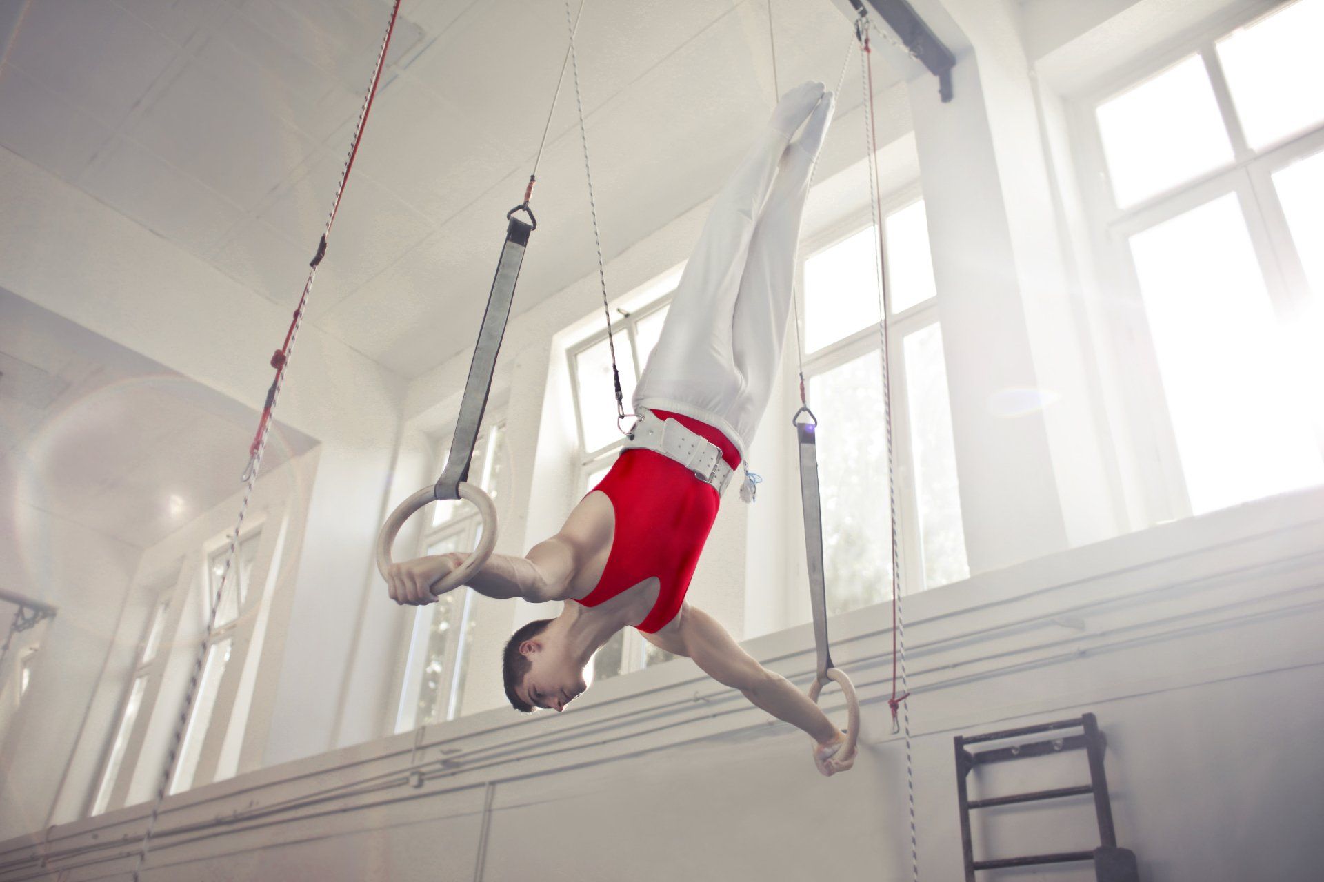 Gymnast in red and white uniform performing on rings in a bright, indoor gym.