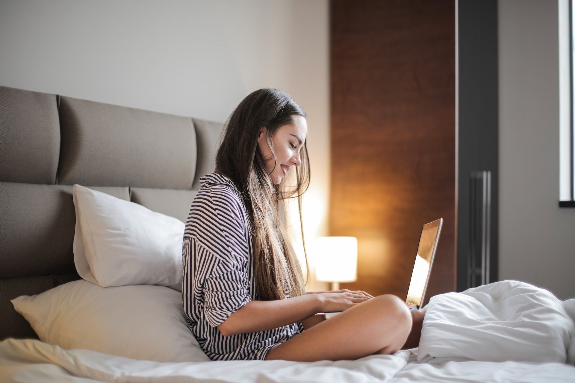 Eine Frau sitzt lächelnd auf einem Bett und benutzt einen Laptop. Warmes Licht erhellt den Raum.