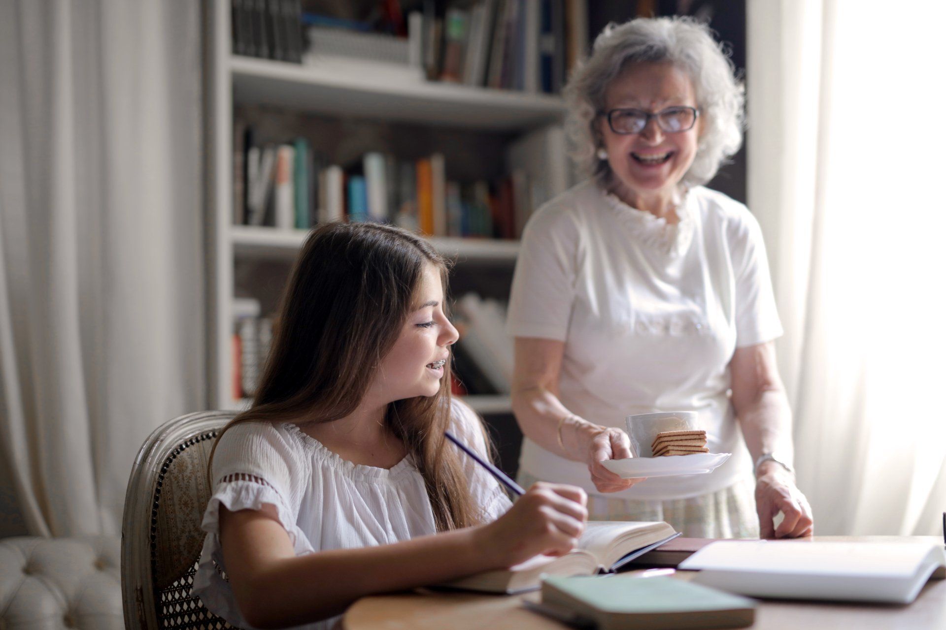 A person writing in a book at a table while someone offers them a plate with a snack, in a room with a bookshelf.