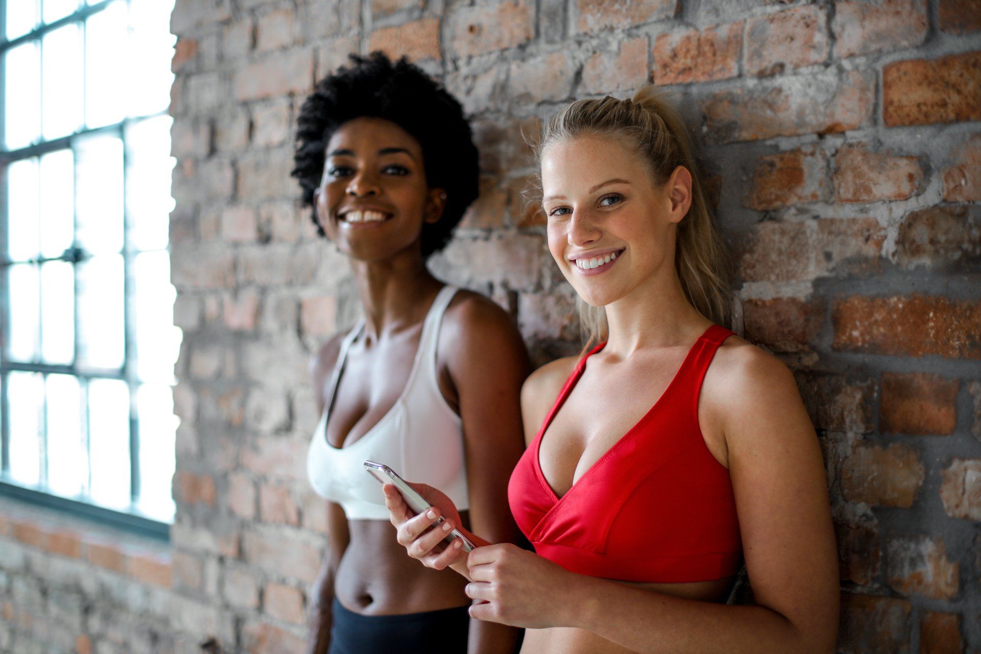 Two women in workout tops smile by a brick wall, one holding a phone near a bright window.