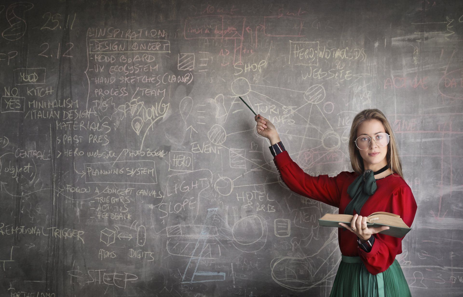 A woman is standing in front of a blackboard holding a book to teach and provide marriage and family therapy supervision.