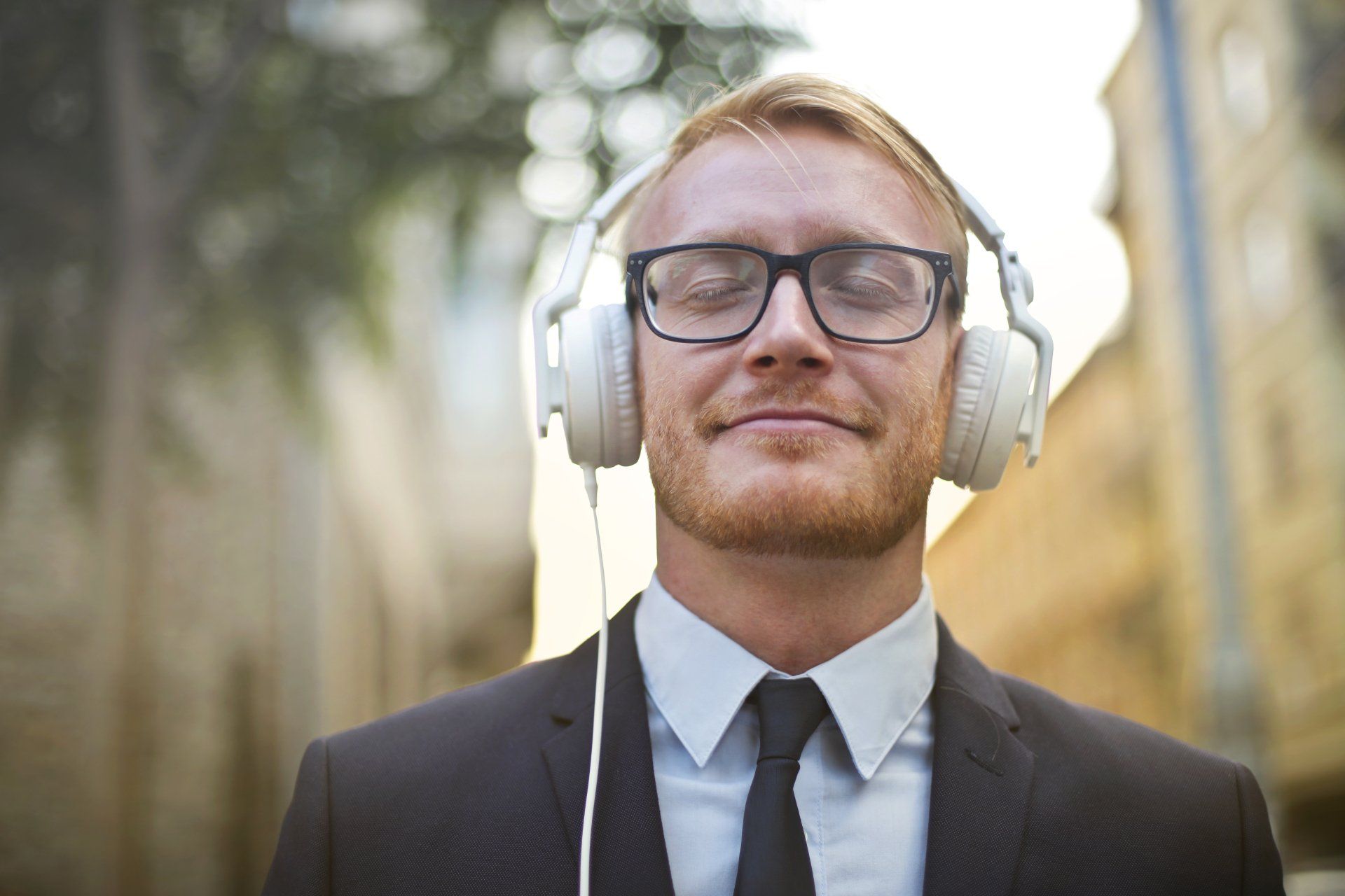 Un homme en costume, lunettes et casque audio, les yeux fermés, sourit à l'extérieur.