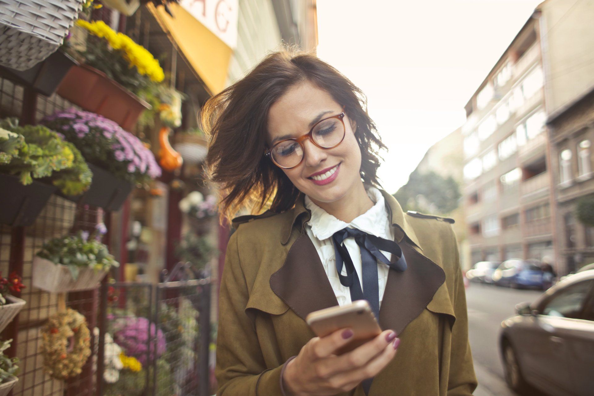 Smiling woman in glasses looking at her phone on a city street beside flower displays