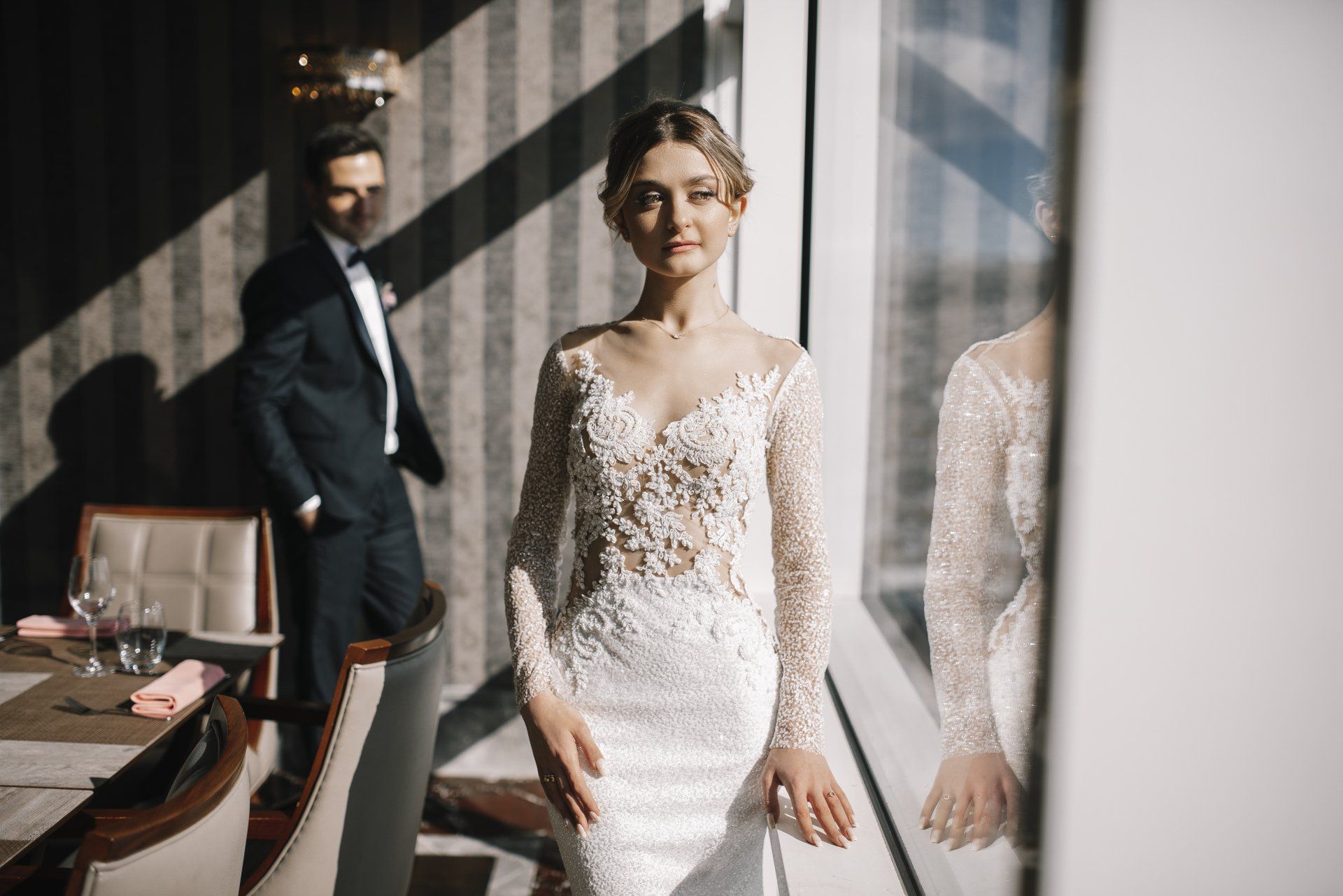 A bride and groom are standing next to each other in front of a window.