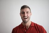 Man with short brown hair and beard smiles broadly, wearing a red polo shirt, against a white background.