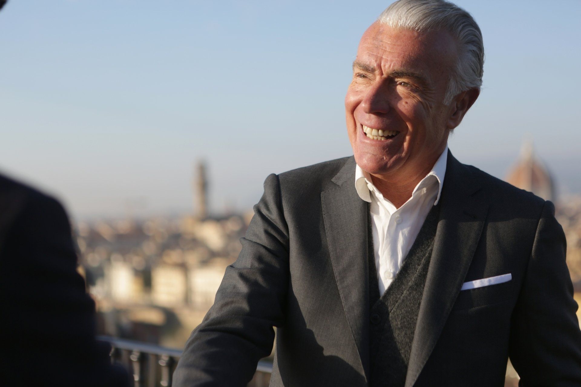 Smiling man in a dark suit outdoors with a city skyline in the background