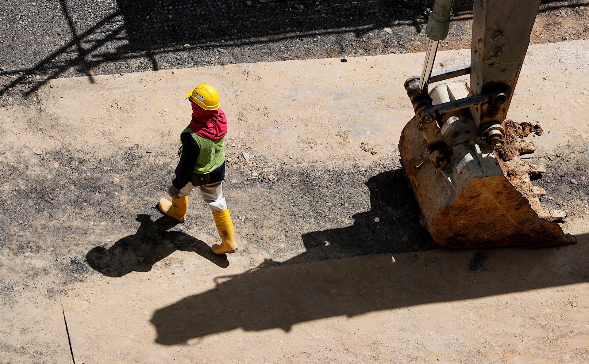 Construction worker in yellow gear walking beside a large excavator bucket on a dirt site.