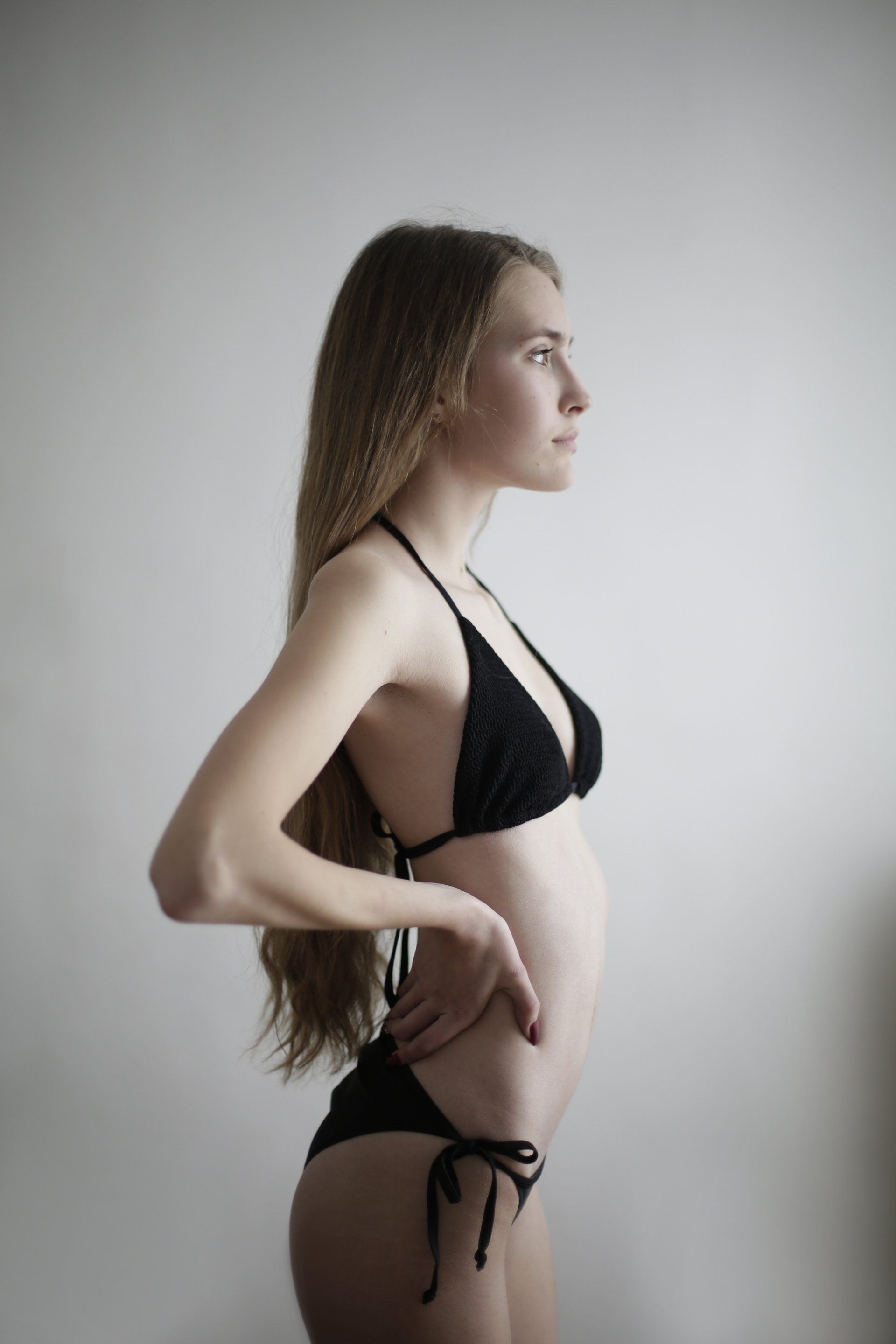 Mujer en bikini negro, de perfil lateral, posando con la mano en la cadera, contra una pared blanca.