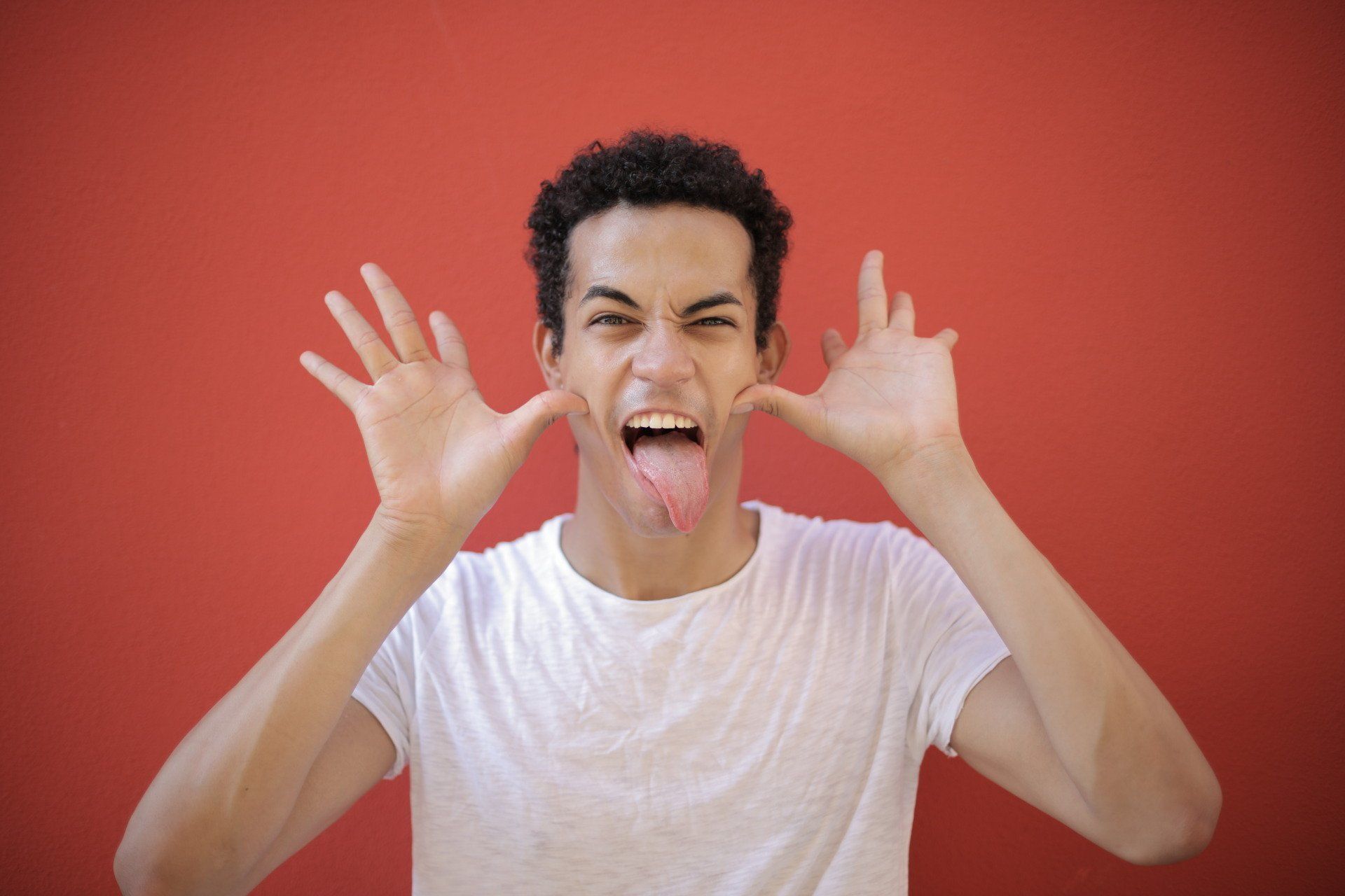 Person with a white t-shirt making a face, sticking out tongue and spreading fingers, against a red background.