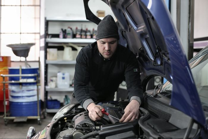 Mechanic in a garage working on a car engine with a screwdriver, wearing a black beanie.