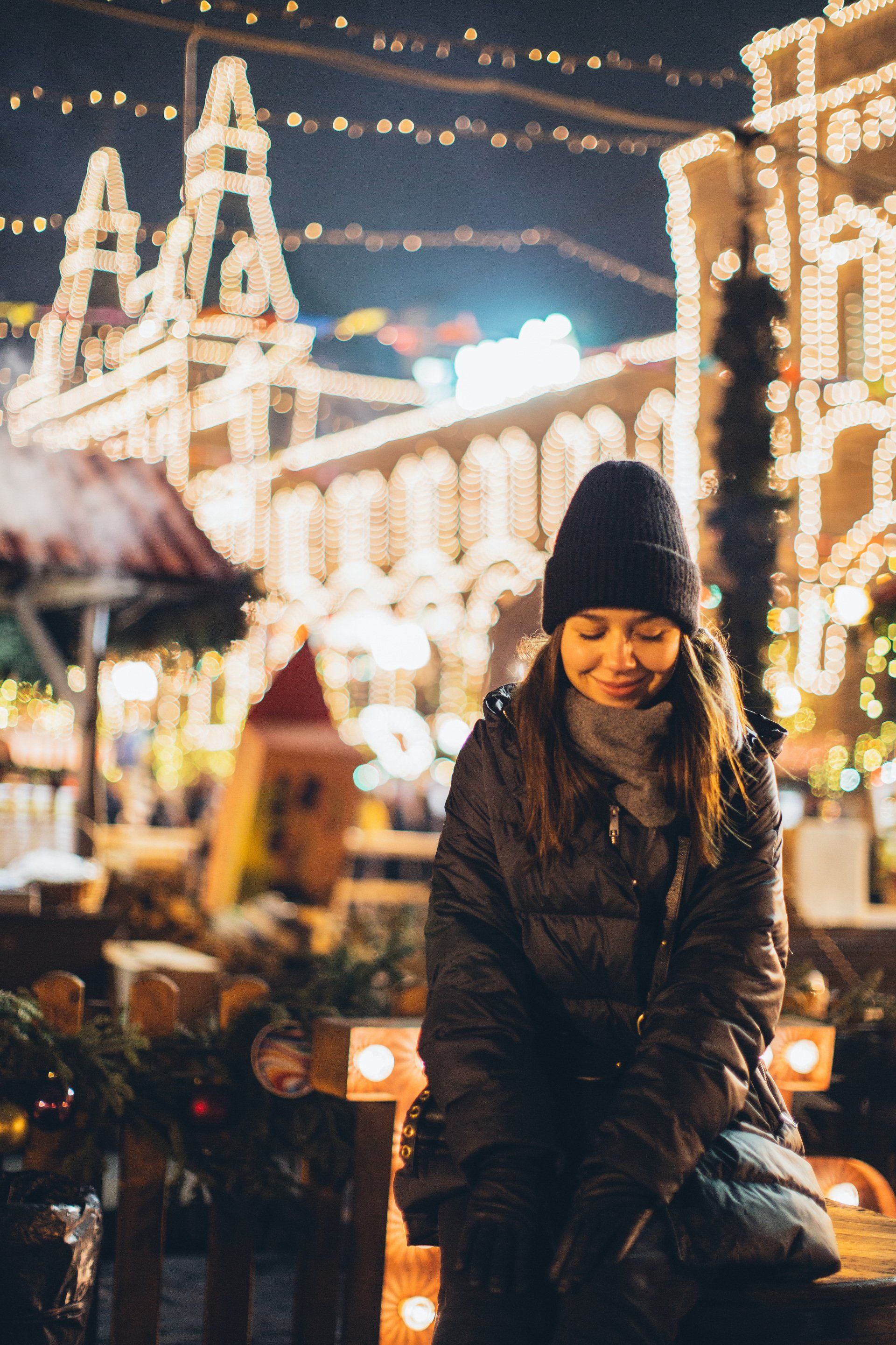 Femme emmitouflée dans un manteau et un bonnet d'hiver, souriante, illuminée par les lumières de Noël.