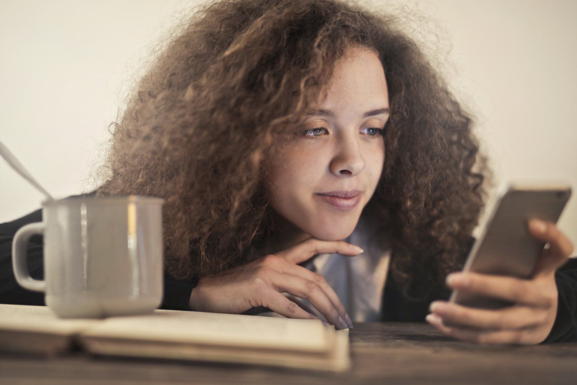 Woman with curly hair looking at a phone, leaning on a wooden table with a mug and open book.