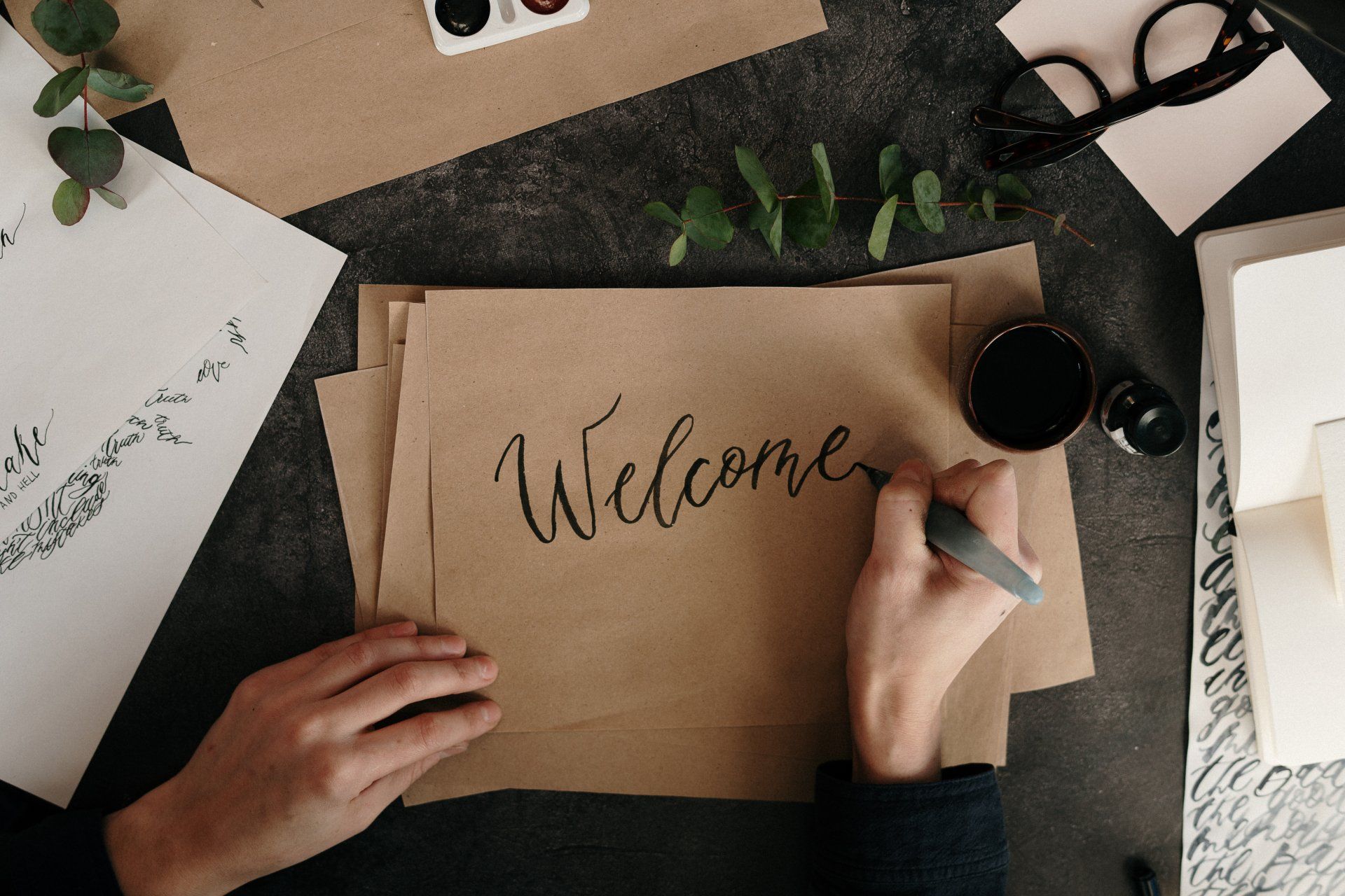 photo of hands with right hand writing the word Welcome on a brown stack of papers