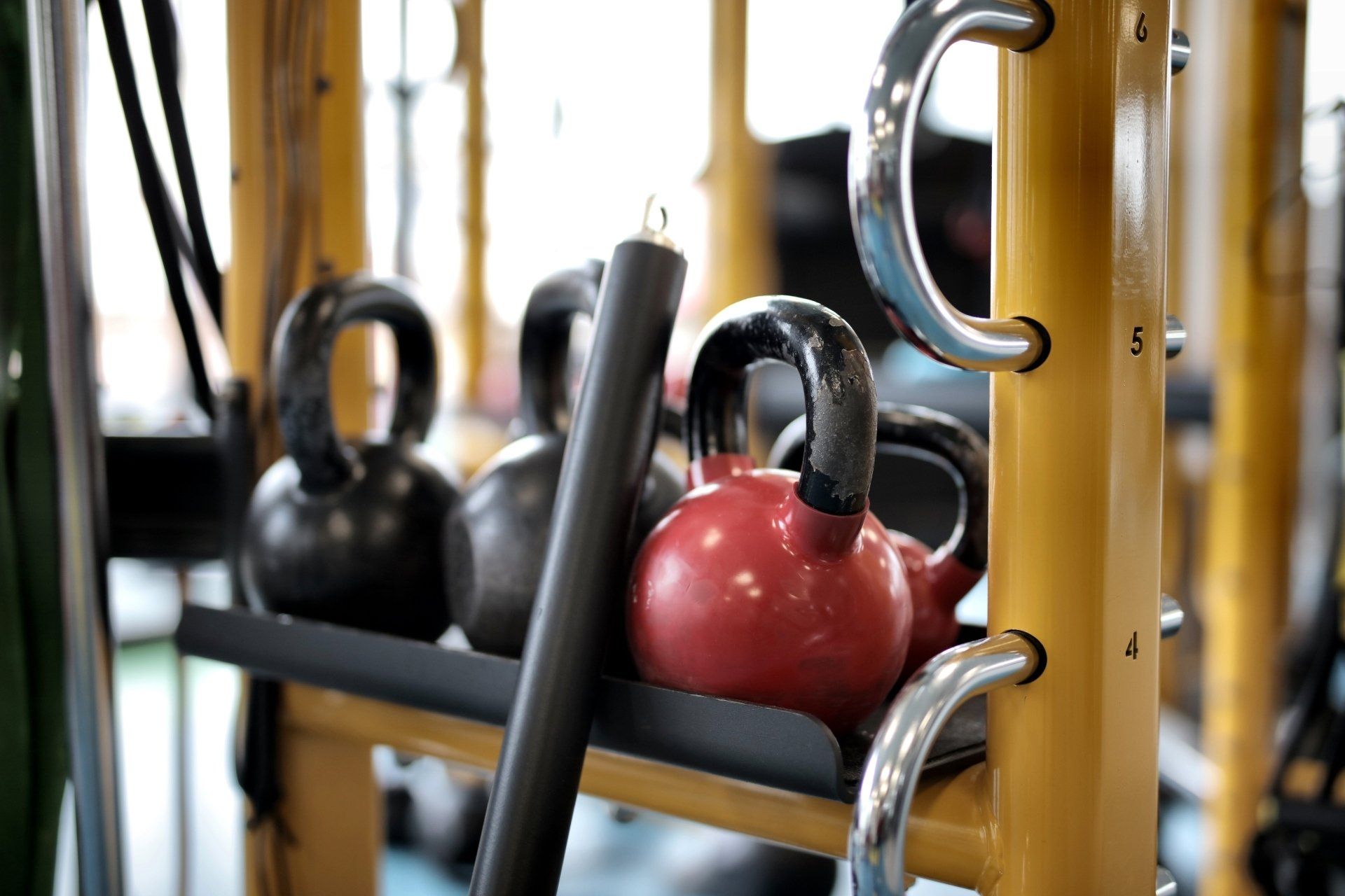 Kettlebells on a weight rack in a gym setting. Red, black, and other weights are visible with metal handles.