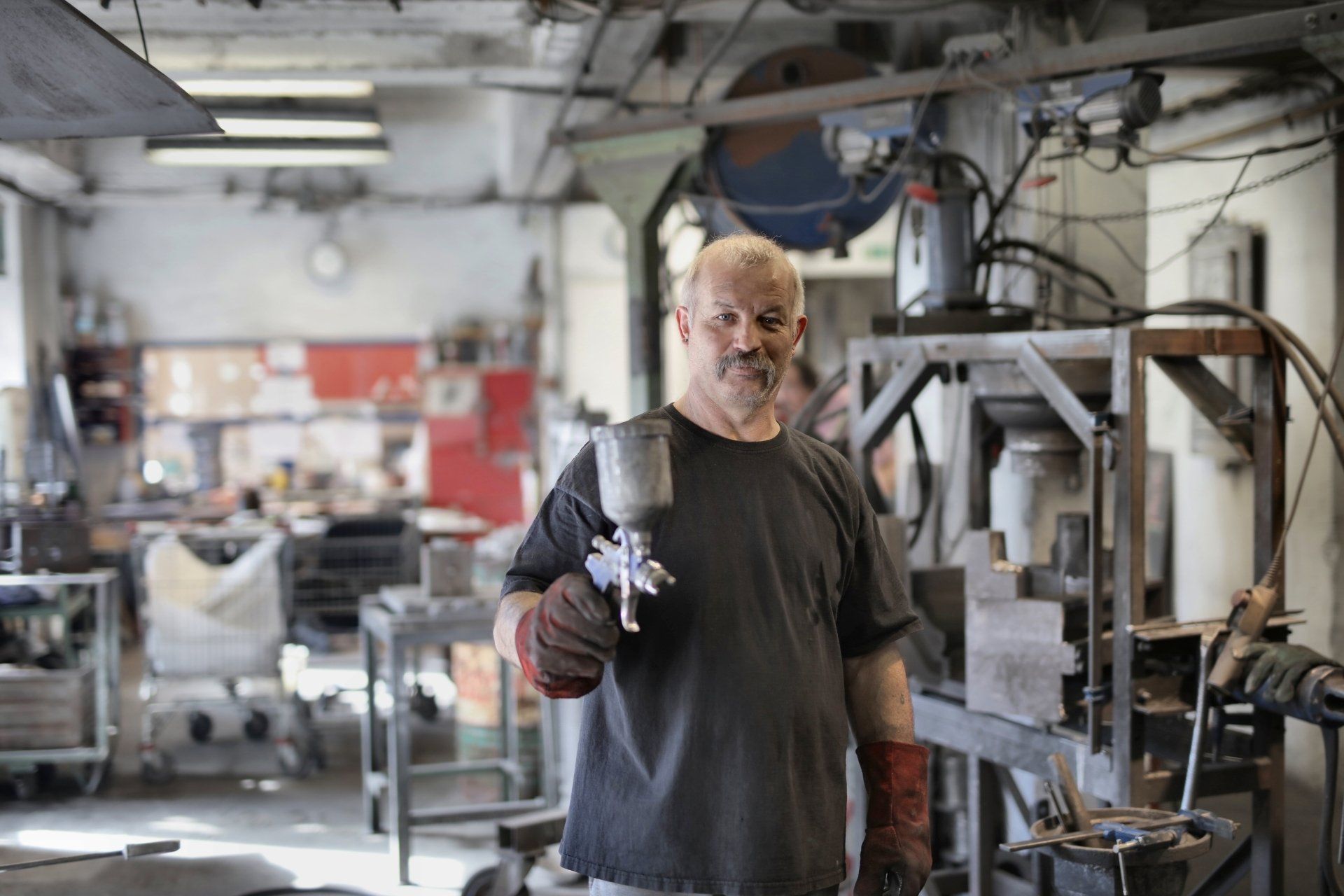 Un homme dans un atelier tient un pistolet à peinture, porte des gants et sourit.