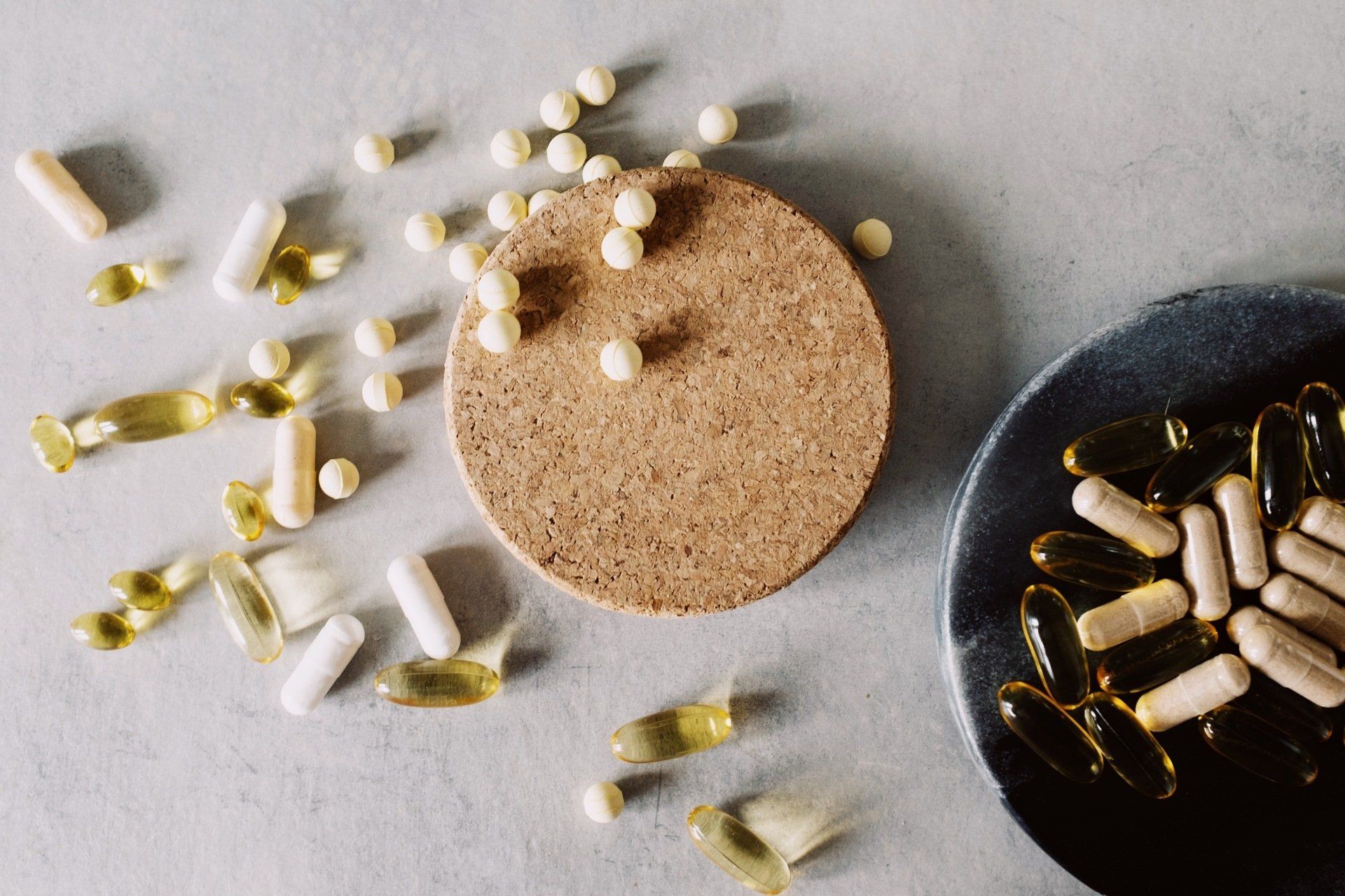 Assorted pills and capsules scattered around cork and black bowls on a gray surface