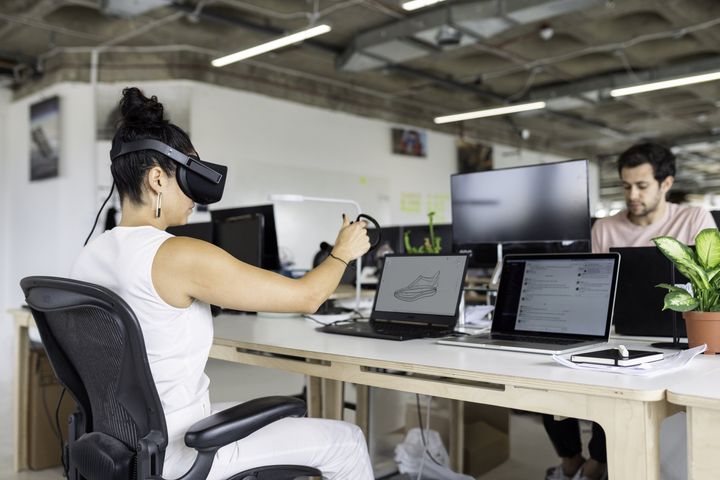 A woman wearing a VR headset at a desk, holding a controller. A man works at a computer nearby in an office.