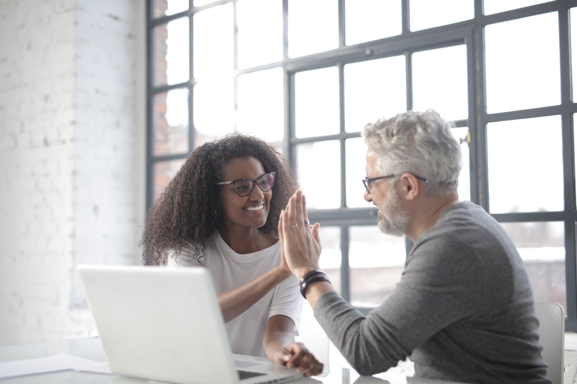 A man and a woman are giving each other a high five while sitting at a table with a laptop.