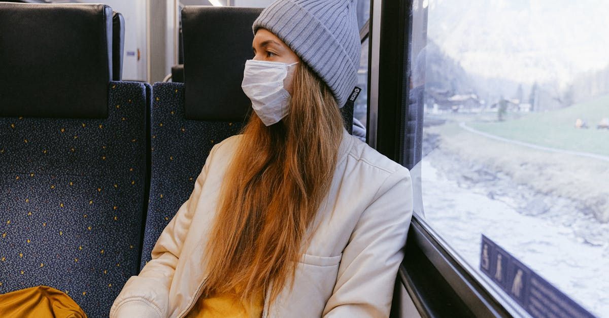 A person wearing a grey beanie and a white face mask sits on a train, looking out a window at a snowy landscape.