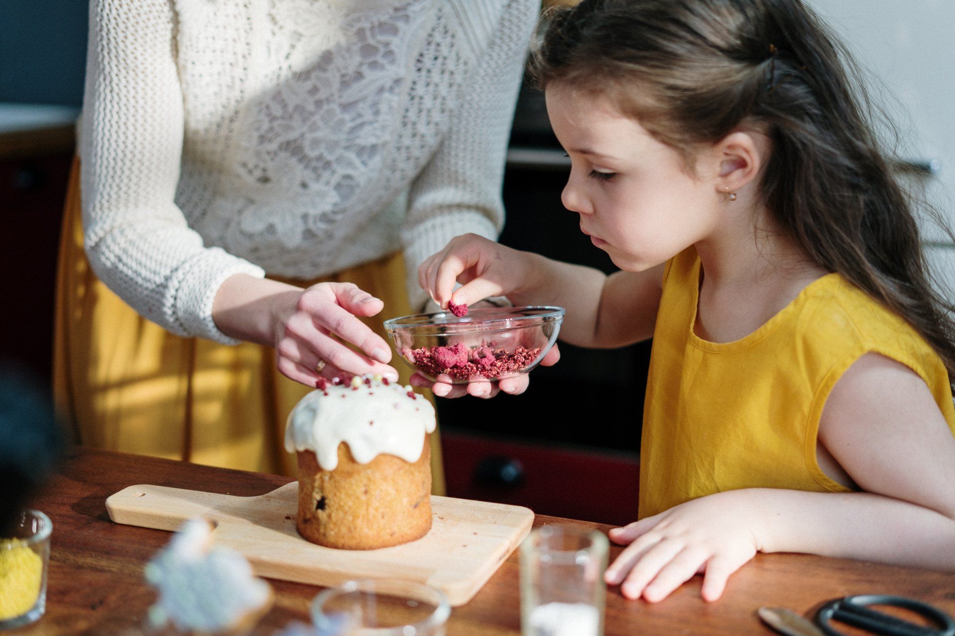 Adult and child decorating a cake with pink frosting and cherries at a wooden table