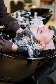 Woman getting hair washed in a salon sink; face relaxed, eyes closed, surrounded by suds.