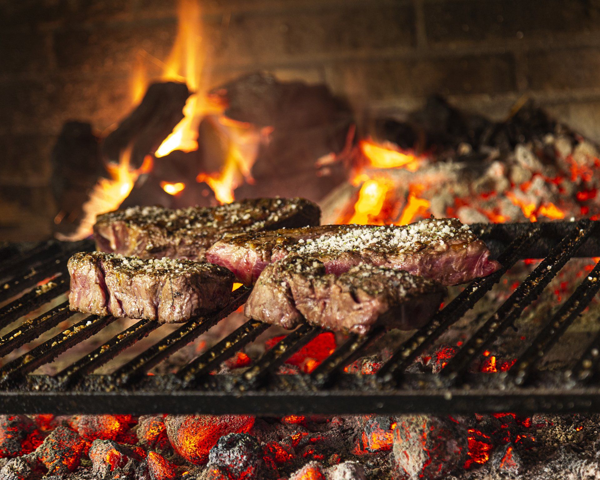 Steaks grilling over hot coals in an outdoor fireplace; flames in the background.
