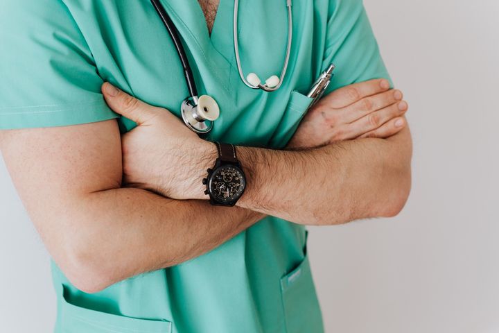 Un uomo con un camice verde e uno stetoscopio al collo è in piedi con le braccia incrociate.