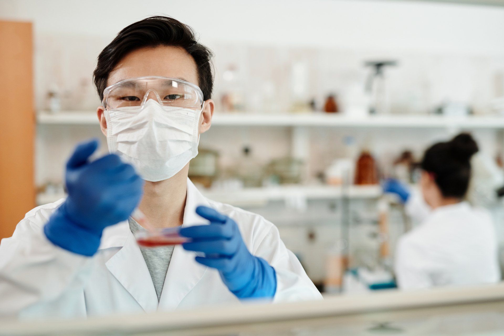 A scientist wearing a mask and goggles is holding a petri dish in a laboratory.