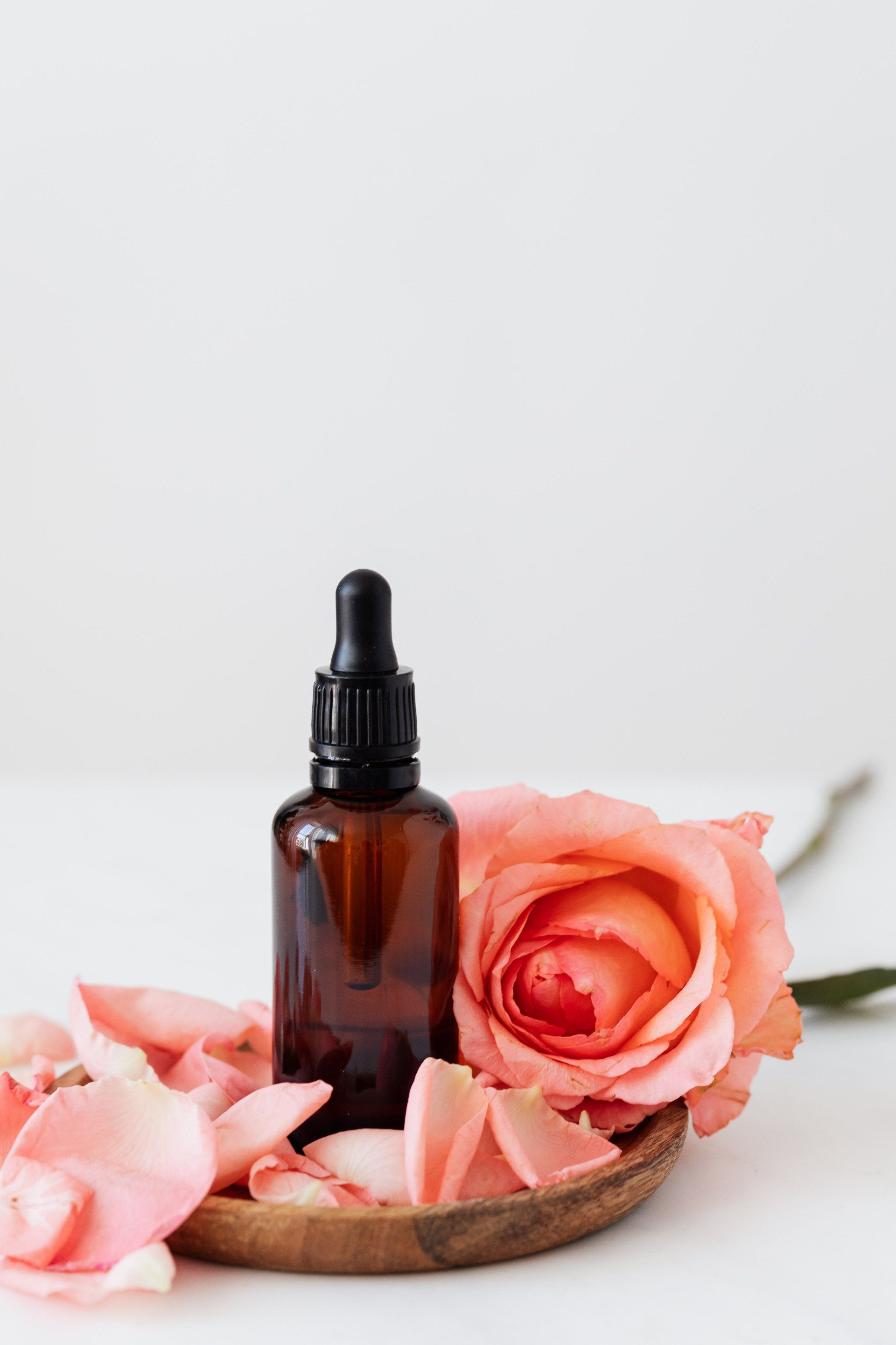 Una bottiglia di olio essenziale accanto a una rosa rosa e petali su un piatto di legno.