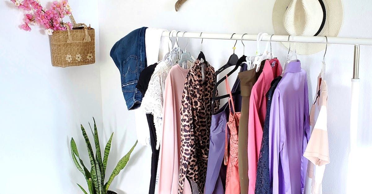 Aan een wit rek hangen kleren; roze, paarse en bruine kledingstukken. Aan de muur hangt een gevlochten mand met roze bloemen.
