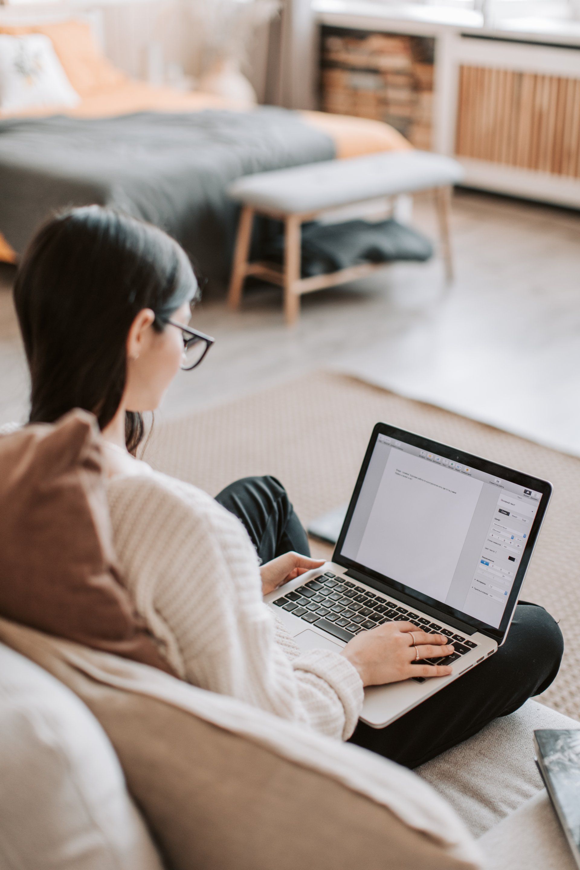 A person in a light-colored sweater sits on a couch and types on a laptop in a bright, modern room.