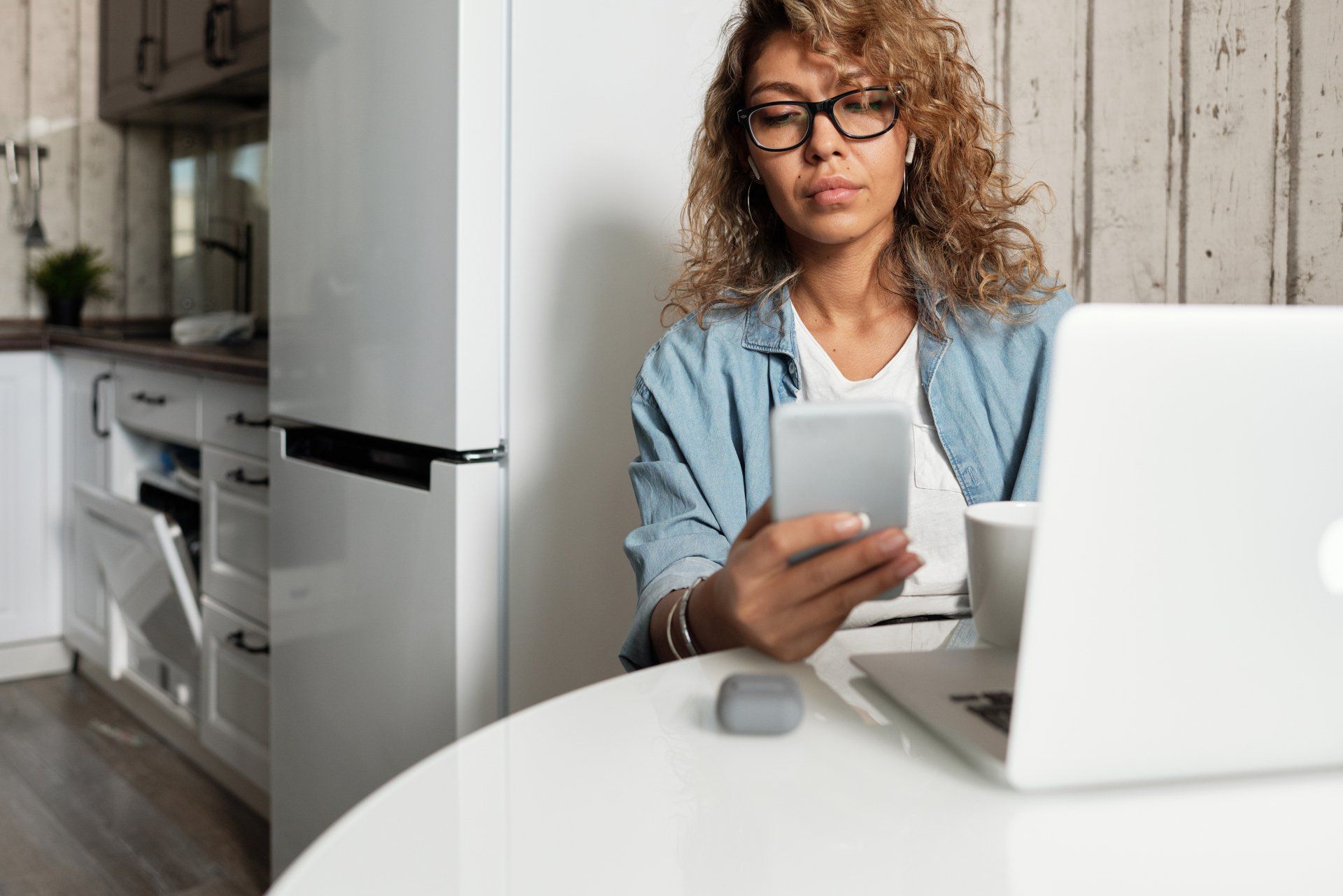 A person with curly hair wearing glasses sits at a kitchen table, looking at a smartphone next to an open laptop.