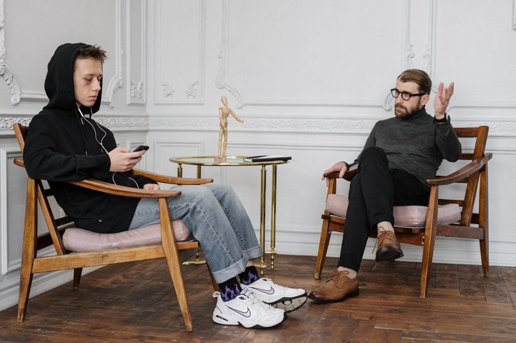 A person in a hoodie and jeans sits, looking at their phone, while a person in glasses gestures in a room with wood floors.
