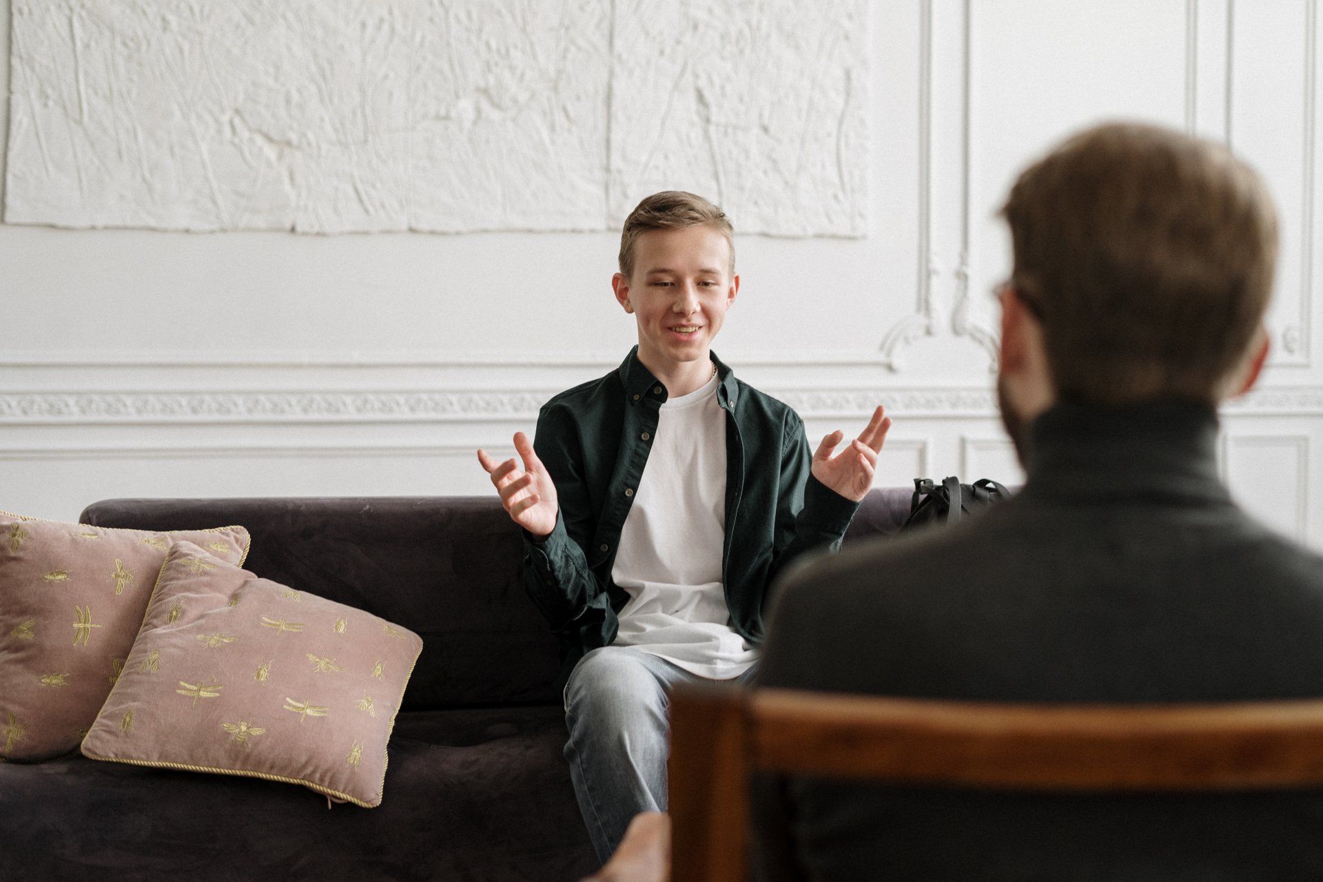 Man on couch talking to person in chair, gesturing with hands. White room, neutral decor.