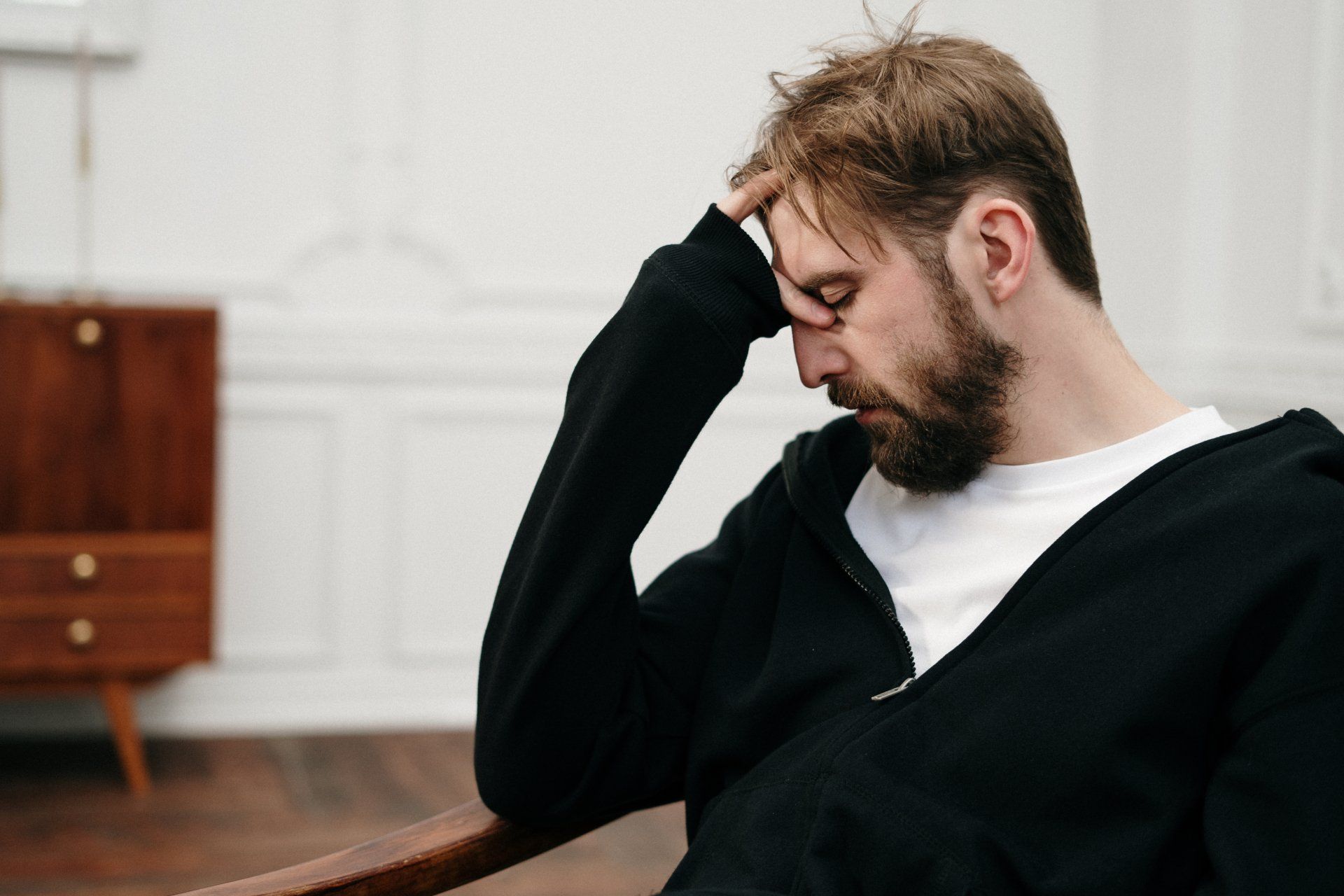 Man in a black sweater sits with his head in his hand, looking stressed in a bright room