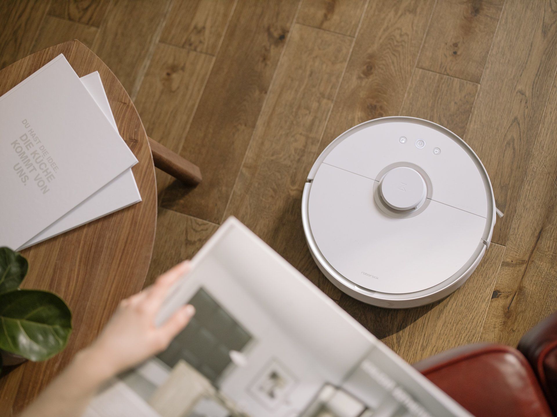 A person is reading a magazine next to a robot vacuum cleaner.