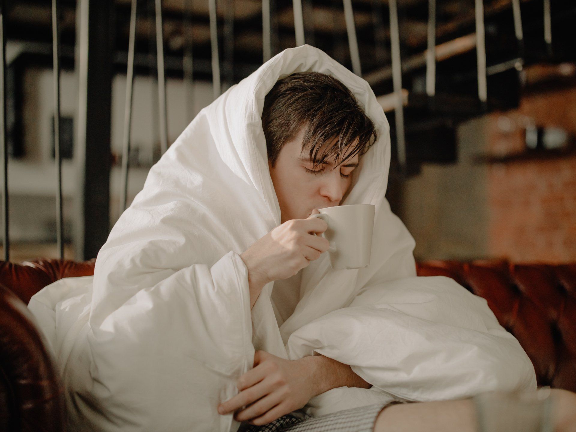 A man wrapped in a blanket is sitting on a couch drinking a cup of coffee.