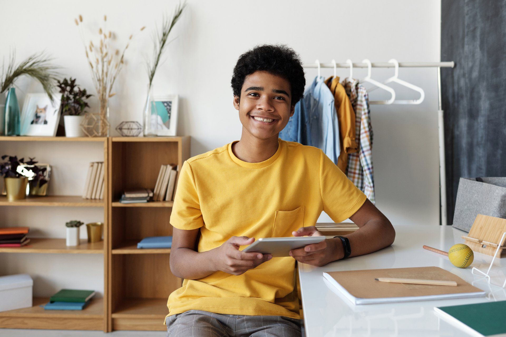 Person smiling at a desk in a bright home office, holding a tablet with shelves and decor behind them